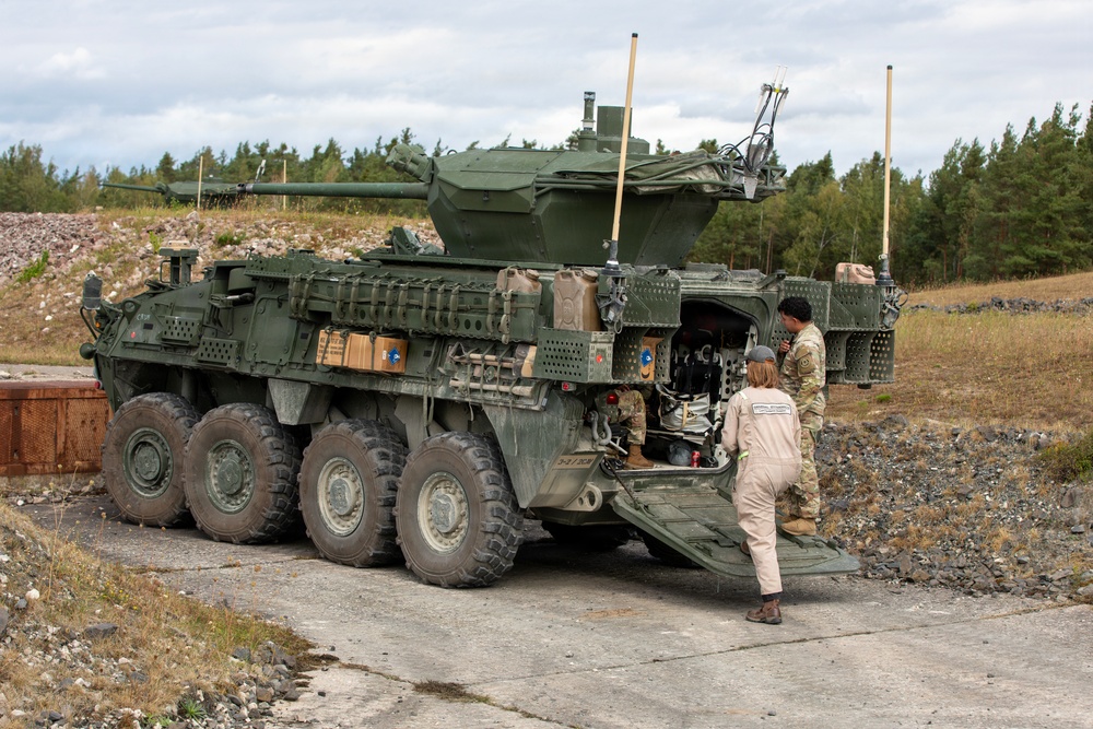 A large 8-wheeled armored military vehicle, consistent with a Stryker Infantry Carrier Vehicle (ICV) fitted with a remote weapon station turret, is parked on a concrete surface in what appears to be a training or exercise area. Three individuals are visible near the rear ramp of the vehicle — one in military camouflage uniform, one in a tan/beige civilian or contractor jumpsuit, and one seated inside the vehicle. The setting includes a rocky terrain, sparse dry grass, and a treeline of conifers under an overcast sky.