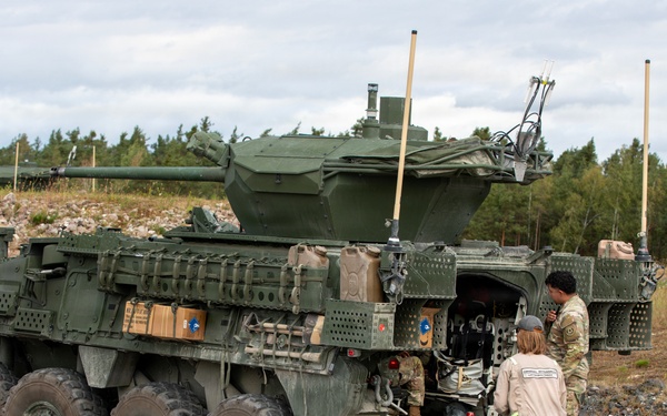 A large 8-wheeled armored military vehicle, consistent with a Stryker Infantry Carrier Vehicle (ICV) fitted with a remote weapon station turret, is parked on a concrete surface in what appears to be a training or exercise area. Three individuals are visible near the rear ramp of the vehicle — one in military camouflage uniform, one in a tan/beige civilian or contractor jumpsuit, and one seated inside the vehicle. The setting includes a rocky terrain, sparse dry grass, and a treeline of conifers under an overcast sky.