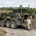 A large 8-wheeled armored military vehicle, consistent with a Stryker Infantry Carrier Vehicle (ICV) fitted with a remote weapon station turret, is parked on a concrete surface in what appears to be a training or exercise area. Three individuals are visible near the rear ramp of the vehicle — one in military camouflage uniform, one in a tan/beige civilian or contractor jumpsuit, and one seated inside the vehicle. The setting includes a rocky terrain, sparse dry grass, and a treeline of conifers under an overcast sky.