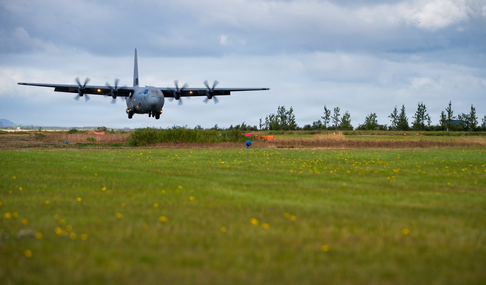 A military turboprop transport aircraft, consistent with a C-130 Hercules design, is captured at very low altitude during an approach or low-level pass over a grass field. The aircraft's four propellers are visibly spinning, and its landing gear appears to be extended. The setting appears to be a rural or semi-improved airstrip with trees and orange marker cones visible in the background.