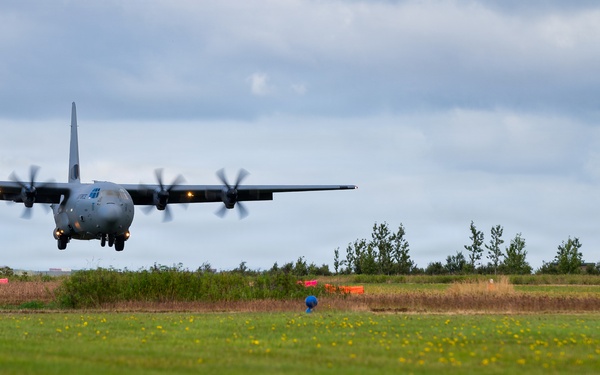 A military turboprop transport aircraft, consistent with a C-130 Hercules design, is captured at very low altitude during an approach or low-level pass over a grass field. The aircraft's four propellers are visibly spinning, and its landing gear appears to be extended. The setting appears to be a rural or semi-improved airstrip with trees and orange marker cones visible in the background.