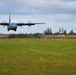A military turboprop transport aircraft, consistent with a C-130 Hercules design, is captured at very low altitude during an approach or low-level pass over a grass field. The aircraft's four propellers are visibly spinning, and its landing gear appears to be extended. The setting appears to be a rural or semi-improved airstrip with trees and orange marker cones visible in the background.