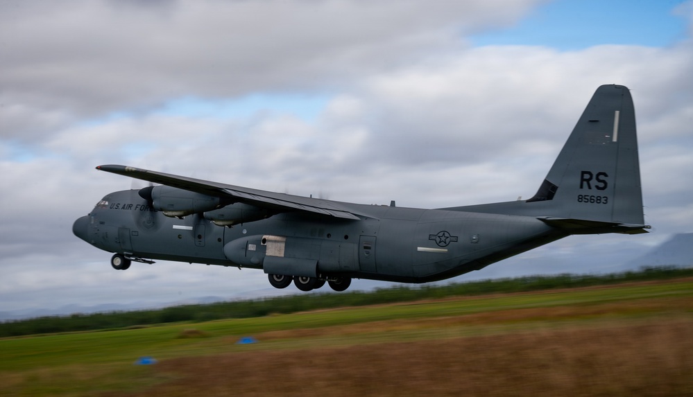 A U.S. Air Force C-130 Hercules military transport aircraft is captured in low-altitude flight, likely during takeoff or landing. The aircraft bears tail code 'RS' and serial number '85683', painted in overall gray livery. Motion blur on the ground and vegetation indicates the photo was taken with a panning technique to track the moving aircraft.