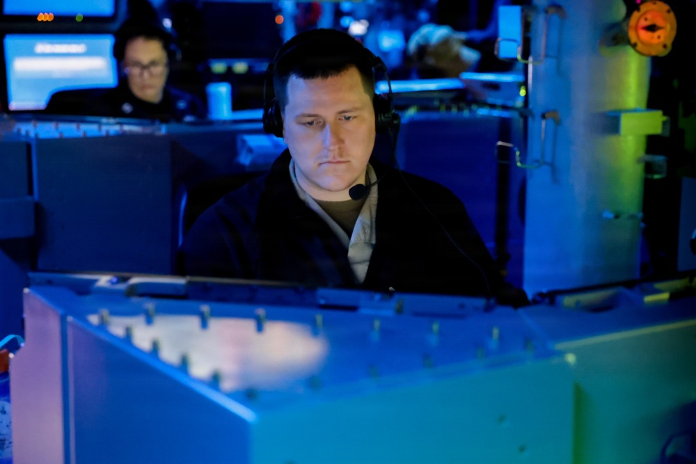 A person wearing a headset with a boom microphone is seated at what appears to be a military or naval operations console, illuminated by blue ambient lighting. A second individual wearing glasses is visible in the background, also seated at a workstation with monitors. The environment suggests a command-and-control or combat information center setting.