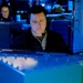 A person wearing a headset with a boom microphone is seated at what appears to be a military or naval operations console, illuminated by blue ambient lighting. A second individual wearing glasses is visible in the background, also seated at a workstation with monitors. The environment suggests a command-and-control or combat information center setting.