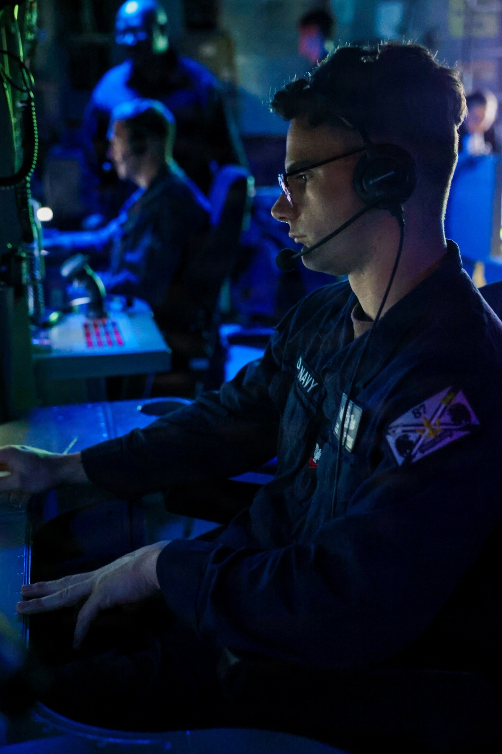 A U.S. Navy sailor is seated at a workstation in a dimly lit combat information center or operations room, wearing a headset with boom microphone and glasses. The environment is illuminated primarily by blue ambient lighting, consistent with a shipboard tactical operations space. Additional personnel are visible in the background at other workstations.