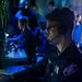 A U.S. Navy sailor is seated at a workstation in a dimly lit combat information center or operations room, wearing a headset with boom microphone and glasses. The environment is illuminated primarily by blue ambient lighting, consistent with a shipboard tactical operations space. Additional personnel are visible in the background at other workstations.
