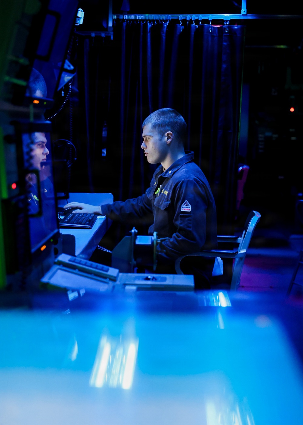 A young military personnel member, wearing a dark navy uniform with a visible patch, is seated at a workstation operating a keyboard in a dimly lit operations or command center. A second individual is partially visible in a monitor or reflective surface to the left. The environment is characterized by blue ambient lighting, dark curtains, and multiple electronic displays, consistent with a naval combat information center or bridge.