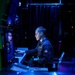 A young military personnel member, wearing a dark navy uniform with a visible patch, is seated at a workstation operating a keyboard in a dimly lit operations or command center. A second individual is partially visible in a monitor or reflective surface to the left. The environment is characterized by blue ambient lighting, dark curtains, and multiple electronic displays, consistent with a naval combat information center or bridge.