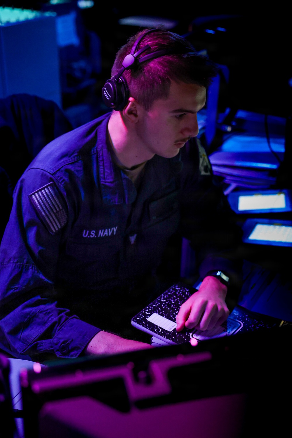A U.S. Navy sailor is seated at a workstation in a dimly lit operations or combat information center, wearing headphones and a navy blue uniform. The sailor is focused on tasks at the console, holding what appears to be a composition notebook. The environment is illuminated by blue and purple ambient lighting typical of shipboard tactical spaces.