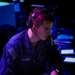 A U.S. Navy sailor is seated at a workstation in a dimly lit operations or combat information center, wearing headphones and a navy blue uniform. The sailor is focused on tasks at the console, holding what appears to be a composition notebook. The environment is illuminated by blue and purple ambient lighting typical of shipboard tactical spaces.