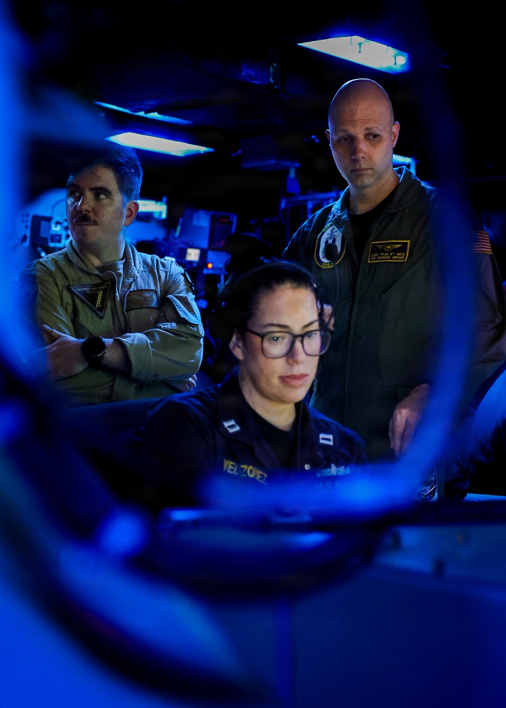 Three military personnel are visible in what appears to be a darkened combat information center (CIC) or operations room aboard a naval vessel. A seated female sailor wearing glasses and a name tag reading 'VELAZQUEZ' is focused on a monitor, while two male officers in flight suits stand behind her observing. The scene is illuminated primarily by blue ambient lighting and screen glow, suggesting an active operational environment.