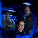 Three military personnel are visible in what appears to be a darkened combat information center (CIC) or operations room aboard a naval vessel. A seated female sailor wearing glasses and a name tag reading 'VELAZQUEZ' is focused on a monitor, while two male officers in flight suits stand behind her observing. The scene is illuminated primarily by blue ambient lighting and screen glow, suggesting an active operational environment.