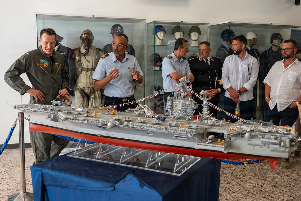 A group of military and civilian personnel are gathered around a highly detailed scale model of an aircraft carrier, numbered '74', displayed on a table in what appears to be a military museum. The model is decorated with small international signal flags and features intricate deck details including miniature aircraft and equipment. Behind the group, a display case contains military helmets, flight suits, and uniforms on mannequins.