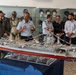 A group of military and civilian personnel are gathered around a highly detailed scale model of an aircraft carrier, numbered '74', displayed on a table in what appears to be a military museum. The model is decorated with small international signal flags and features intricate deck details including miniature aircraft and equipment. Behind the group, a display case contains military helmets, flight suits, and uniforms on mannequins.