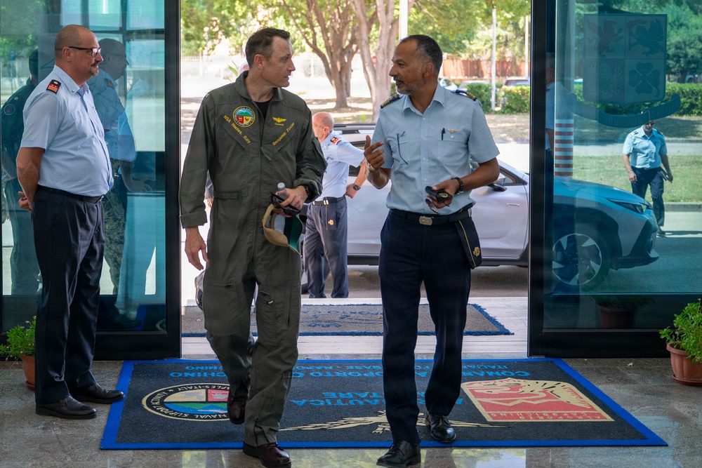 The image shows a meeting or greeting between military personnel at what appears to be a NATO or joint military installation, likely in Italy based on the floor mat text referencing 'Naval Support Activity' and Italian language text. A person in an olive green flight suit is conversing with an officer in a light blue uniform with dark trousers, suggesting an inter-service or multinational military exchange. Several other uniformed personnel are visible in the background near glass entrance doors.