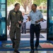 The image shows a meeting or greeting between military personnel at what appears to be a NATO or joint military installation, likely in Italy based on the floor mat text referencing 'Naval Support Activity' and Italian language text. A person in an olive green flight suit is conversing with an officer in a light blue uniform with dark trousers, suggesting an inter-service or multinational military exchange. Several other uniformed personnel are visible in the background near glass entrance doors.