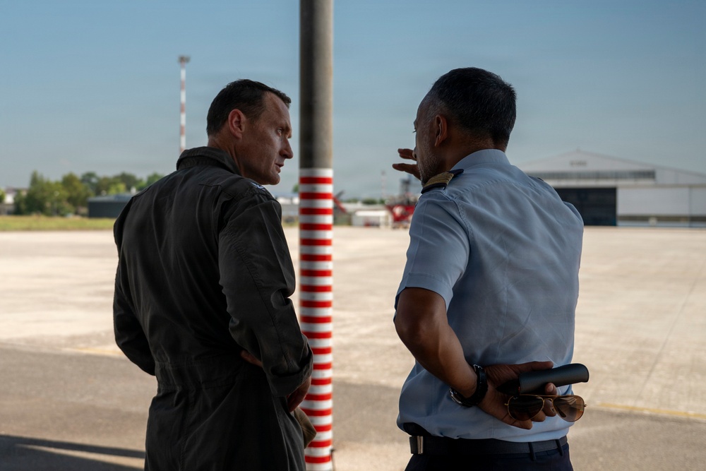 Two men in aviation-related attire are engaged in conversation on an airport tarmac or apron area. One wears a dark flight suit while the other wears a light blue uniform shirt with shoulder epaulettes, suggesting military or aviation authority roles. A red-and-white striped safety pole is visible between them, with aircraft hangars visible in the background.