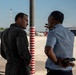 Two men in aviation-related attire are engaged in conversation on an airport tarmac or apron area. One wears a dark flight suit while the other wears a light blue uniform shirt with shoulder epaulettes, suggesting military or aviation authority roles. A red-and-white striped safety pole is visible between them, with aircraft hangars visible in the background.