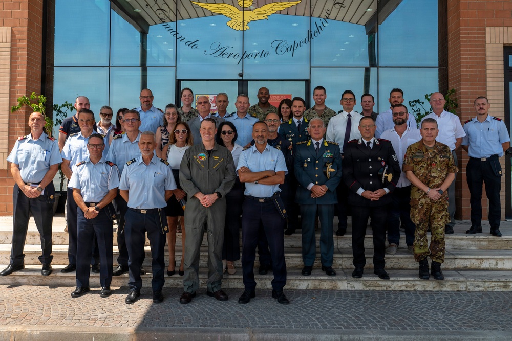 A group photograph of approximately 30 military and civilian personnel posing on the steps of 'Comando Aeroporto Capodichino' (Naples Capodichino Airport Command) in Italy. The group includes members wearing various uniforms representing different branches and nationalities, including Italian Air Force, Carabinieri, Italian Army, and what appears to be US military personnel. The setting suggests an official visit, meeting, or joint military coordination event.