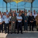 A group photograph of approximately 30 military and civilian personnel posing on the steps of 'Comando Aeroporto Capodichino' (Naples Capodichino Airport Command) in Italy. The group includes members wearing various uniforms representing different branches and nationalities, including Italian Air Force, Carabinieri, Italian Army, and what appears to be US military personnel. The setting suggests an official visit, meeting, or joint military coordination event.