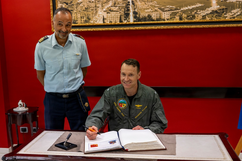 Two military officers are photographed in a formal setting with red walls, where one officer in a green flight suit is signing an official guest book or register at a desk. The standing officer wears a light blue uniform with shoulder epaulettes and a wing badge, suggesting an air force affiliation. The occasion appears to be a formal military visit or ceremony.
