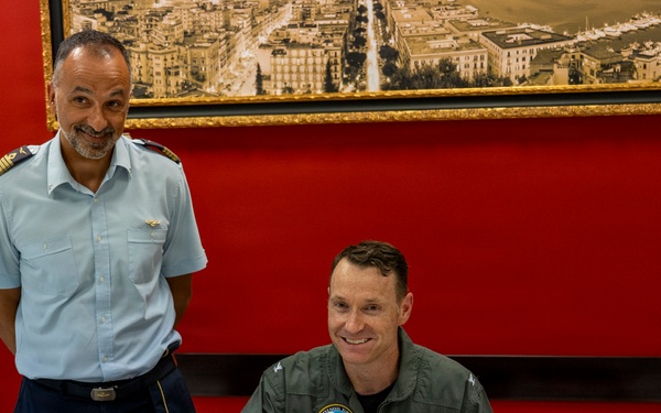 Two military officers are photographed in a formal setting with red walls, where one officer in a green flight suit is signing an official guest book or register at a desk. The standing officer wears a light blue uniform with shoulder epaulettes and a wing badge, suggesting an air force affiliation. The occasion appears to be a formal military visit or ceremony.