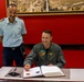 Two military officers are photographed in a formal setting with red walls, where one officer in a green flight suit is signing an official guest book or register at a desk. The standing officer wears a light blue uniform with shoulder epaulettes and a wing badge, suggesting an air force affiliation. The occasion appears to be a formal military visit or ceremony.