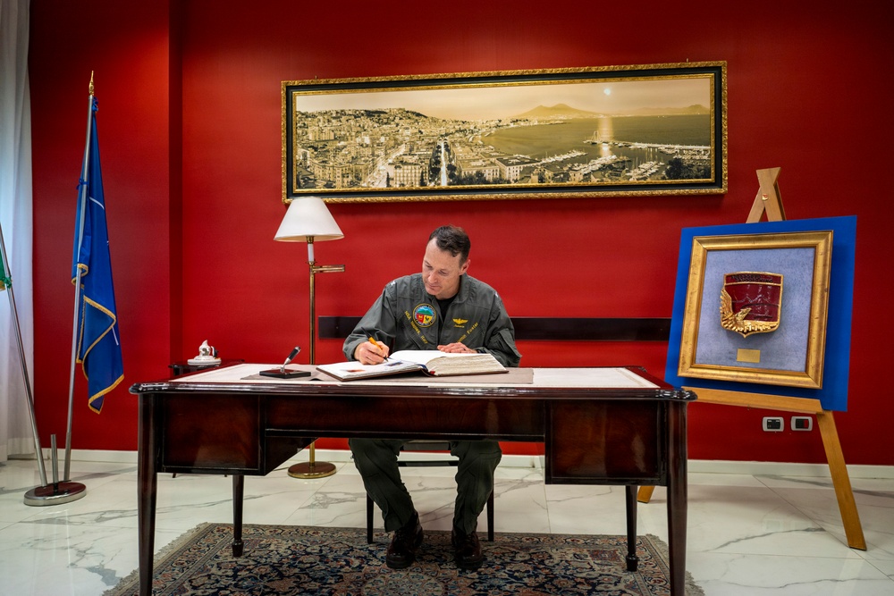 A military officer in an olive/sage green flight suit is seated at a formal mahogany desk, signing or writing in an open book or register. The setting appears to be an official military office with a deep red accent wall, a panoramic cityscape painting above, and a framed military insignia/crest displayed on an easel to the right. The environment suggests a formal ceremonial or administrative occasion.