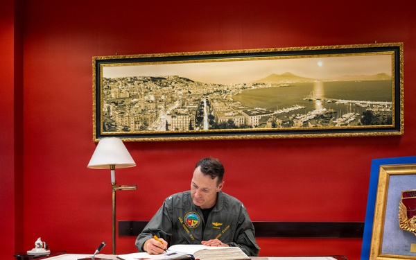 A military officer in an olive/sage green flight suit is seated at a formal mahogany desk, signing or writing in an open book or register. The setting appears to be an official military office with a deep red accent wall, a panoramic cityscape painting above, and a framed military insignia/crest displayed on an easel to the right. The environment suggests a formal ceremonial or administrative occasion.