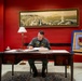 A military officer in an olive/sage green flight suit is seated at a formal mahogany desk, signing or writing in an open book or register. The setting appears to be an official military office with a deep red accent wall, a panoramic cityscape painting above, and a framed military insignia/crest displayed on an easel to the right. The environment suggests a formal ceremonial or administrative occasion.