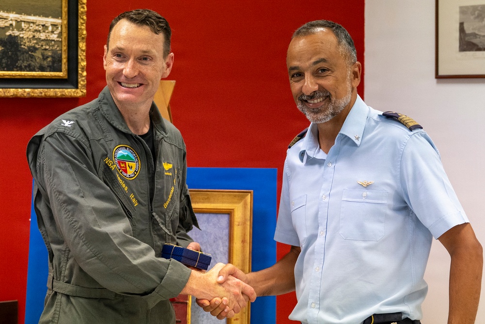 Two military officers are shaking hands in what appears to be an official ceremony or meeting. The officer on the left is wearing a dark olive green flight suit with an NSA Naples, Italy patch, while the officer on the right is wearing a light blue military uniform with rank insignia on the shoulders. They appear to be exchanging or jointly holding a framed item.