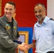 Two military officers are shaking hands in what appears to be an official ceremony or meeting. The officer on the left is wearing a dark olive green flight suit with an NSA Naples, Italy patch, while the officer on the right is wearing a light blue military uniform with rank insignia on the shoulders. They appear to be exchanging or jointly holding a framed item.