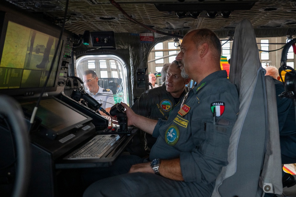 The image shows military personnel inside what appears to be a large aircraft or helicopter cockpit/cabin, examining flight control systems and displays. A man in an Italian military flight suit ('ITALIA' patch visible) is prominently featured alongside colleagues reviewing equipment at a workstation with monitors and a keyboard. The setting suggests a demonstration, briefing, or familiarization tour of the aircraft's mission systems.