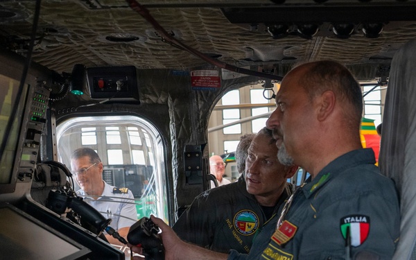 The image shows military personnel inside what appears to be a large aircraft or helicopter cockpit/cabin, examining flight control systems and displays. A man in an Italian military flight suit ('ITALIA' patch visible) is prominently featured alongside colleagues reviewing equipment at a workstation with monitors and a keyboard. The setting suggests a demonstration, briefing, or familiarization tour of the aircraft's mission systems.