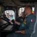 The image shows military personnel inside what appears to be a large aircraft or helicopter cockpit/cabin, examining flight control systems and displays. A man in an Italian military flight suit ('ITALIA' patch visible) is prominently featured alongside colleagues reviewing equipment at a workstation with monitors and a keyboard. The setting suggests a demonstration, briefing, or familiarization tour of the aircraft's mission systems.