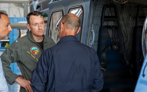 Three military or aviation personnel are gathered near an open helicopter in what appears to be a hangar facility. The central figure wearing an olive green flight suit with a patch is engaged in conversation with the other two individuals. An Italian flag stripe is visible on the helicopter's fuselage, suggesting an Italian military or law enforcement context.