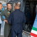 Three military or aviation personnel are gathered near an open helicopter in what appears to be a hangar facility. The central figure wearing an olive green flight suit with a patch is engaged in conversation with the other two individuals. An Italian flag stripe is visible on the helicopter's fuselage, suggesting an Italian military or law enforcement context.