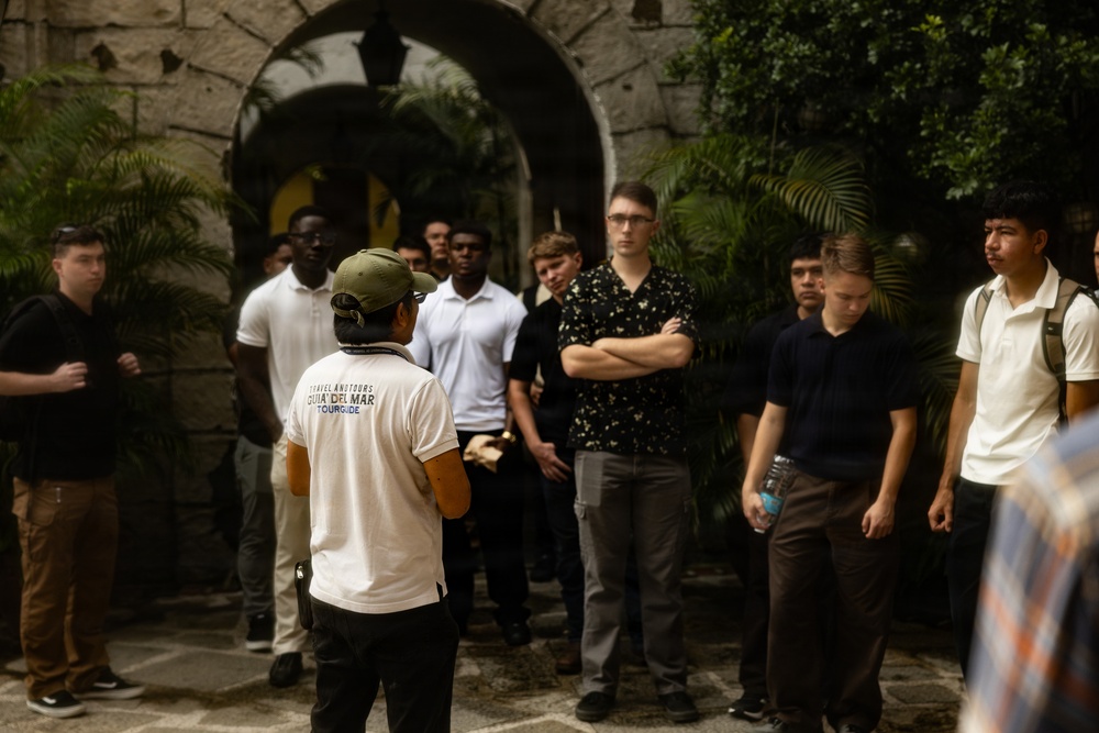 A tour guide wearing a white shirt labeled 'Travel And Tours Guia Del Mar Tour Guide' and a green cap is addressing a group of young men at what appears to be a historic or colonial-style building. The group of approximately 10-12 individuals is gathered in a courtyard or entrance area with stone archways and tropical vegetation. The scene suggests an organized educational or cultural tour.