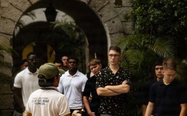 A tour guide wearing a white shirt labeled 'Travel And Tours Guia Del Mar Tour Guide' and a green cap is addressing a group of young men at what appears to be a historic or colonial-style building. The group of approximately 10-12 individuals is gathered in a courtyard or entrance area with stone archways and tropical vegetation. The scene suggests an organized educational or cultural tour.