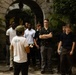 A tour guide wearing a white shirt labeled 'Travel And Tours Guia Del Mar Tour Guide' and a green cap is addressing a group of young men at what appears to be a historic or colonial-style building. The group of approximately 10-12 individuals is gathered in a courtyard or entrance area with stone archways and tropical vegetation. The scene suggests an organized educational or cultural tour.