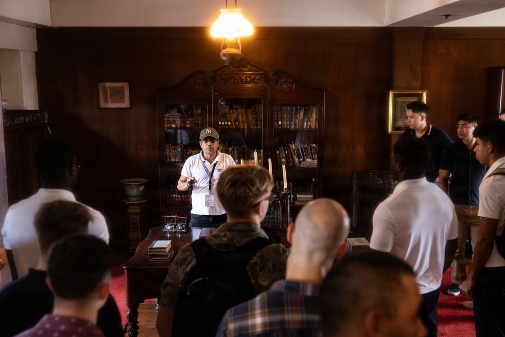 A tour guide wearing a white polo shirt, lanyard, and baseball cap is addressing a group of visitors in what appears to be a historic room or museum study. The room features dark wood paneling, an antique bookcase filled with old books, a wooden desk, and a vintage oil lamp overhead. The guide appears to be gesturing and explaining something to the assembled group.
