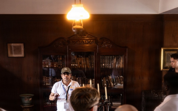 A tour guide wearing a white polo shirt, lanyard, and baseball cap is addressing a group of visitors in what appears to be a historic room or museum study. The room features dark wood paneling, an antique bookcase filled with old books, a wooden desk, and a vintage oil lamp overhead. The guide appears to be gesturing and explaining something to the assembled group.