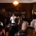 A tour guide wearing a white polo shirt, lanyard, and baseball cap is addressing a group of visitors in what appears to be a historic room or museum study. The room features dark wood paneling, an antique bookcase filled with old books, a wooden desk, and a vintage oil lamp overhead. The guide appears to be gesturing and explaining something to the assembled group.