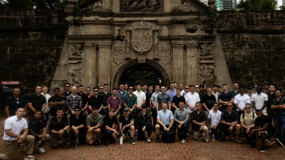 A large group of approximately 50-60 individuals, predominantly men, pose for a group photograph in front of a historic stone gateway featuring an ornate coat of arms relief carving. The structure appears to be Fort Santiago in Intramuros, Manila, Philippines, as indicated by a partially visible sign on the left side. The group is arranged in multiple rows with some kneeling in front and others standing behind.