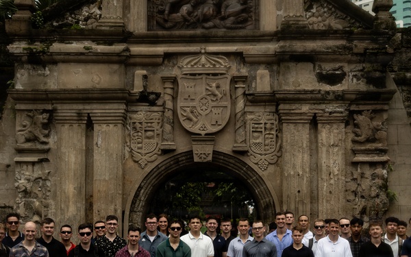 A large group of approximately 50-60 individuals, predominantly men, pose for a group photograph in front of a historic stone gateway featuring an ornate coat of arms relief carving. The structure appears to be Fort Santiago in Intramuros, Manila, Philippines, as indicated by a partially visible sign on the left side. The group is arranged in multiple rows with some kneeling in front and others standing behind.