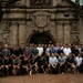 A large group of approximately 50-60 individuals, predominantly men, pose for a group photograph in front of a historic stone gateway featuring an ornate coat of arms relief carving. The structure appears to be Fort Santiago in Intramuros, Manila, Philippines, as indicated by a partially visible sign on the left side. The group is arranged in multiple rows with some kneeling in front and others standing behind.