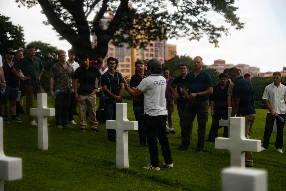 A tour guide wearing a white shirt labeled 'TOURGUIDE' addresses a large group of visitors among white cross grave markers at what appears to be a military cemetery. The setting includes well-maintained green grass, large trees, and urban buildings visible in the background, suggesting a location within or near a city. The group consists predominantly of young adult males listening attentively to the guide's presentation.