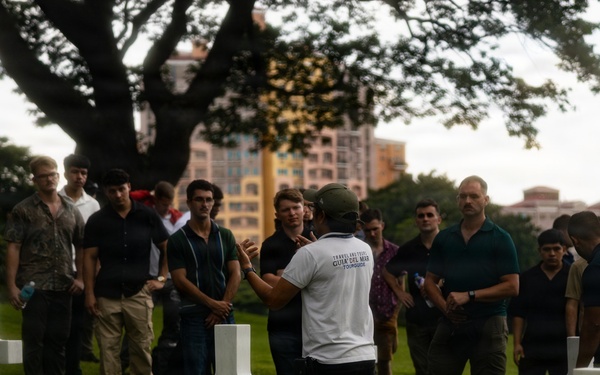 A tour guide wearing a white shirt labeled 'TOURGUIDE' addresses a large group of visitors among white cross grave markers at what appears to be a military cemetery. The setting includes well-maintained green grass, large trees, and urban buildings visible in the background, suggesting a location within or near a city. The group consists predominantly of young adult males listening attentively to the guide's presentation.