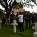 A tour guide wearing a white shirt labeled 'TOURGUIDE' addresses a large group of visitors among white cross grave markers at what appears to be a military cemetery. The setting includes well-maintained green grass, large trees, and urban buildings visible in the background, suggesting a location within or near a city. The group consists predominantly of young adult males listening attentively to the guide's presentation.
